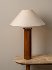 A table lamp with a fluted dark wood column base and a tapered off-white fabric shade, shown lit on a stone-topped table against a warm beige wall.