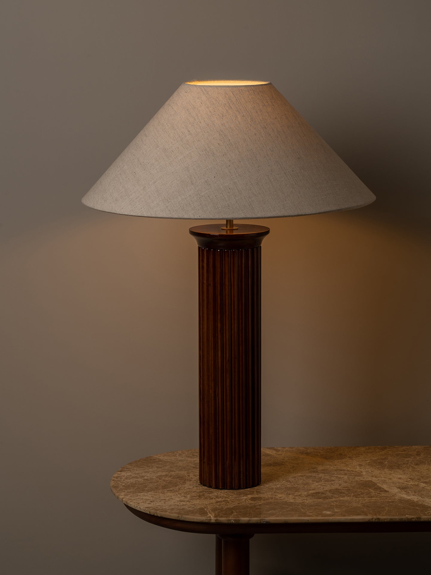 A table lamp with a dark brown fluted column base and a beige fabric drum shade, lit to cast a warm glow against a neutral gray background.