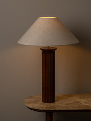A table lamp with a dark brown fluted column base and a beige fabric drum shade, lit to cast a warm glow against a neutral gray background.