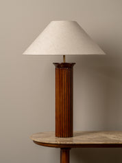 A mid-century modern table lamp with a fluted dark brown wooden column base and a tapered off-white fabric shade, shown on a stone-topped side table against a neutral beige wall.