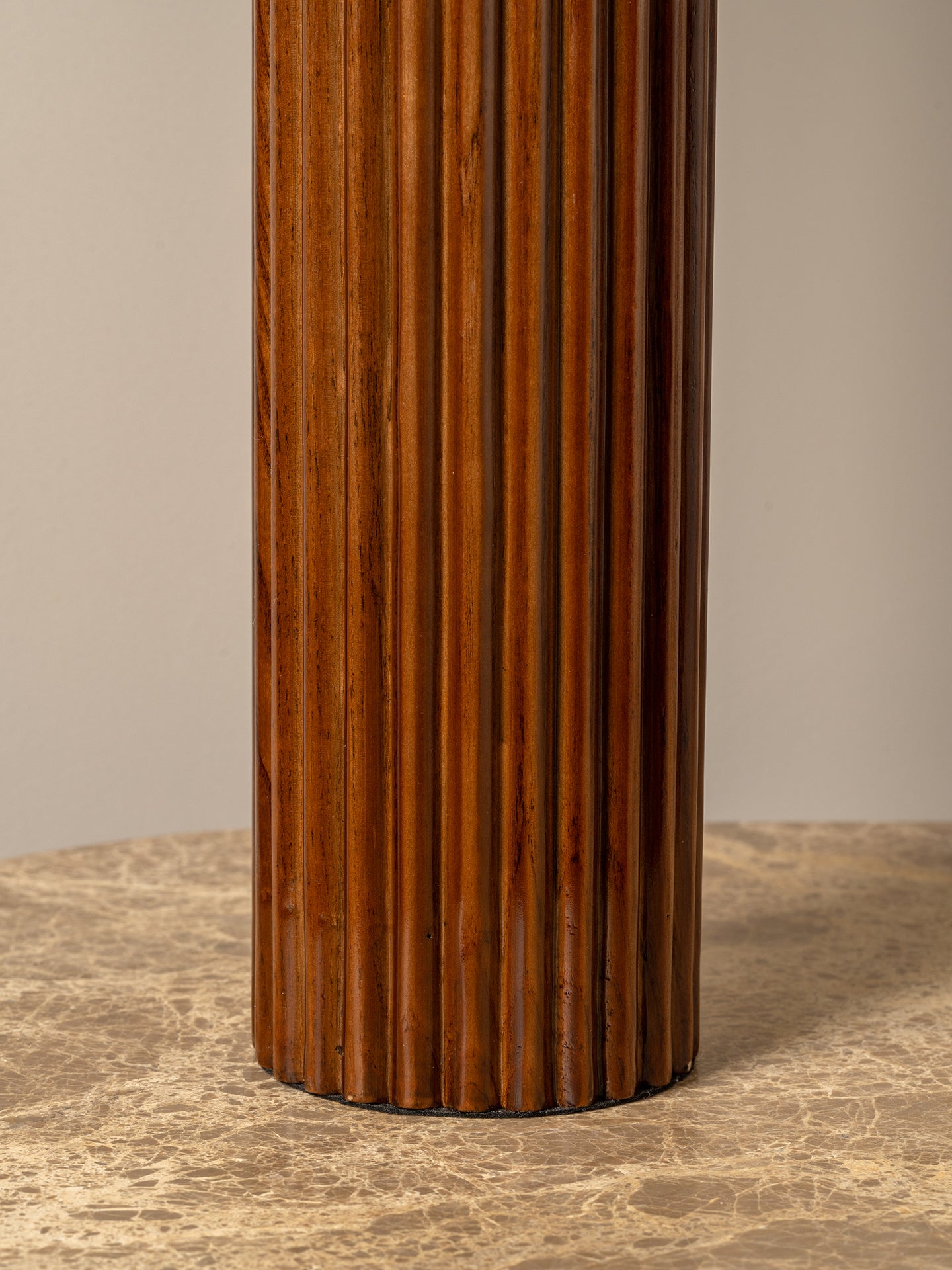 A close-up of a fluted cylindrical column in a warm medium-to-dark brown wood finish, standing on a textured beige stone surface against a neutral beige background.
