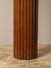 A close-up of a fluted cylindrical column in a warm medium-to-dark brown wood finish, standing on a textured beige stone surface against a neutral beige background.