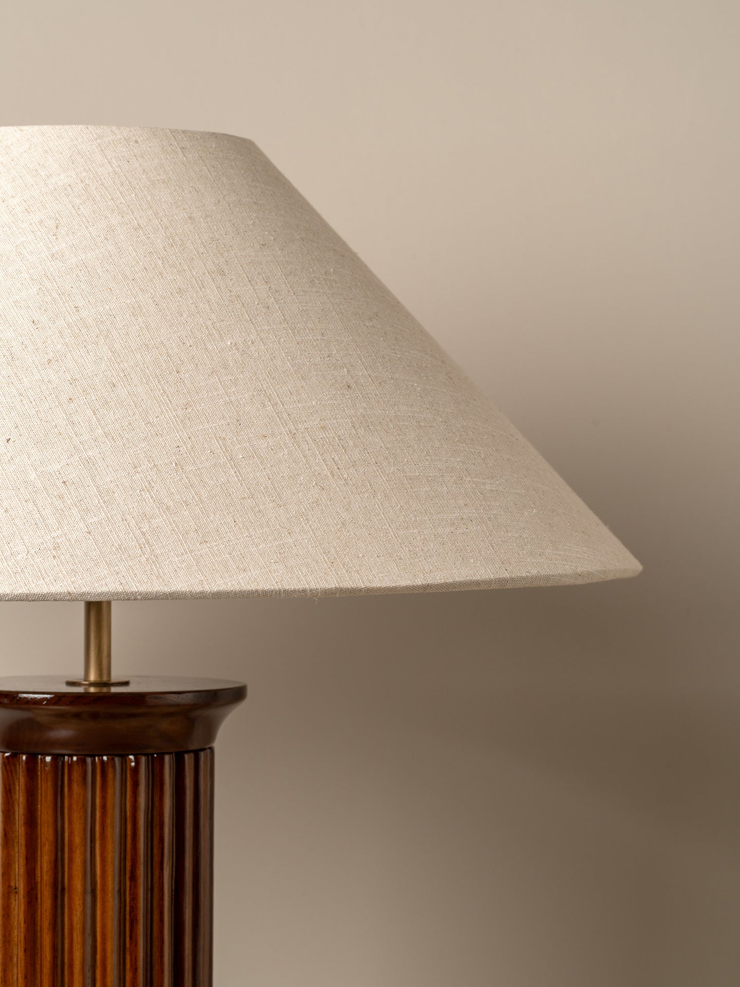 A close-up of a table lamp with a large conical beige linen shade and a dark brown fluted column base, set against a solid warm beige background.