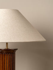 A close-up of a table lamp with a large conical beige linen shade and a dark brown fluted column base, set against a solid warm beige background.