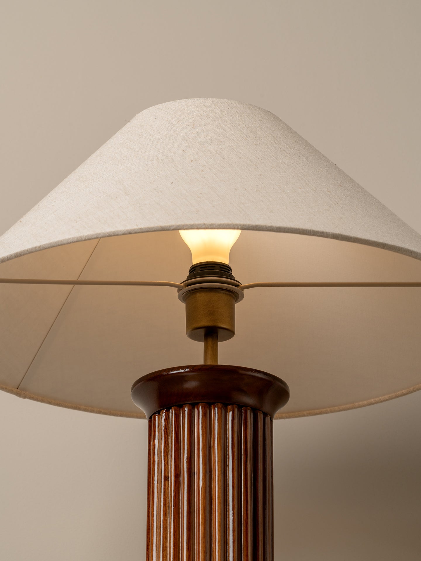 A close-up of a table lamp with a warm glowing light bulb, a beige fabric drum shade, and a fluted dark brown wooden column base.