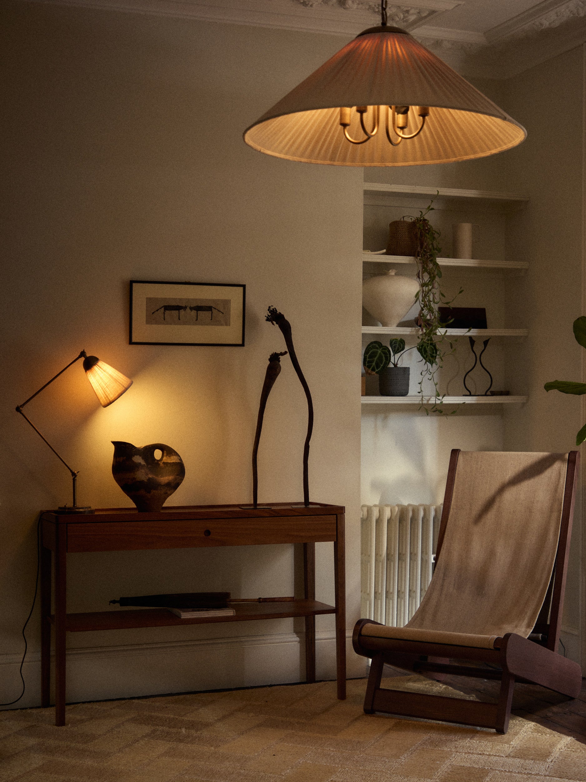 A warm, moody interior scene featuring a large pleated conical pendant lamp hanging from the ceiling, casting a soft glow. Below, a wooden console table holds a lit floor lamp, a dark ceramic vessel, and a sculptural black metal object.