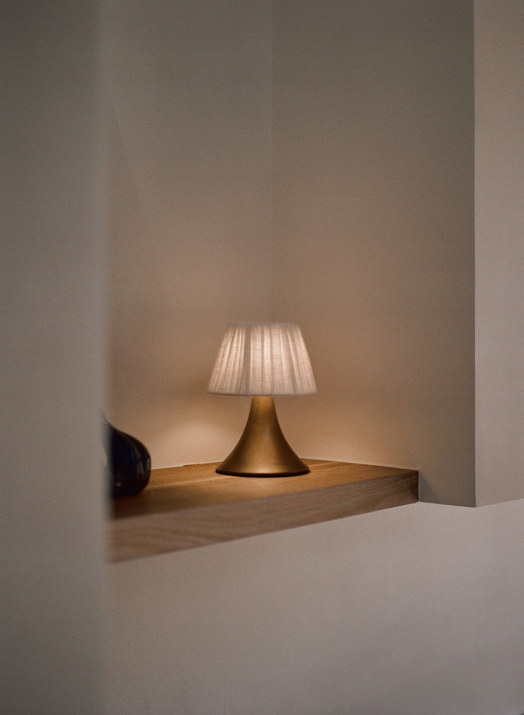A modern table lamp with a pleated white fabric shade and a brushed brass base, illuminated and resting on a light wood shelf against a warm beige wall.