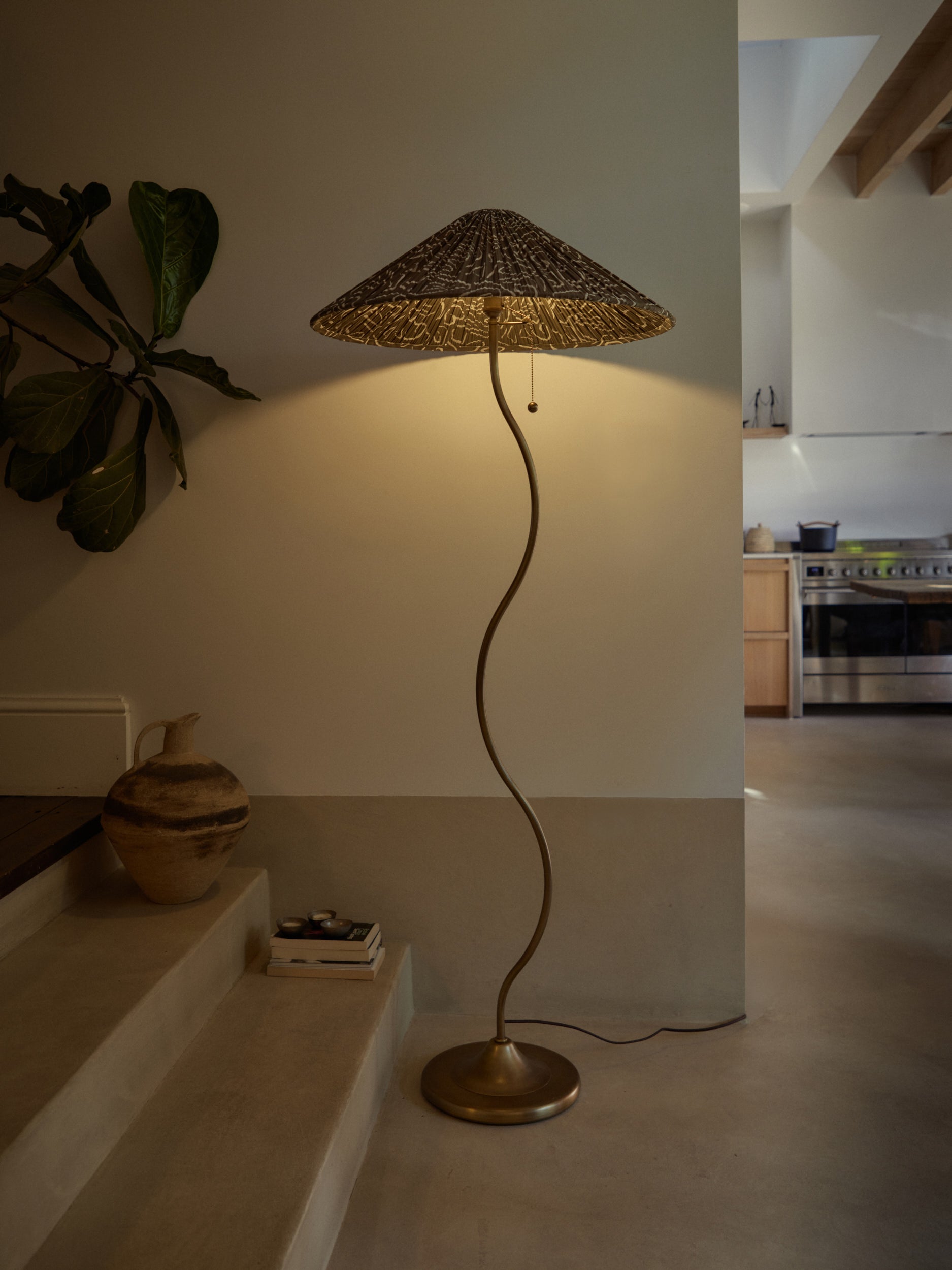 A tall floor lamp with a woven conical shade and a curved metallic stem, shown in a real interior space with stairs, a plant, and a kitchen visible in the background.