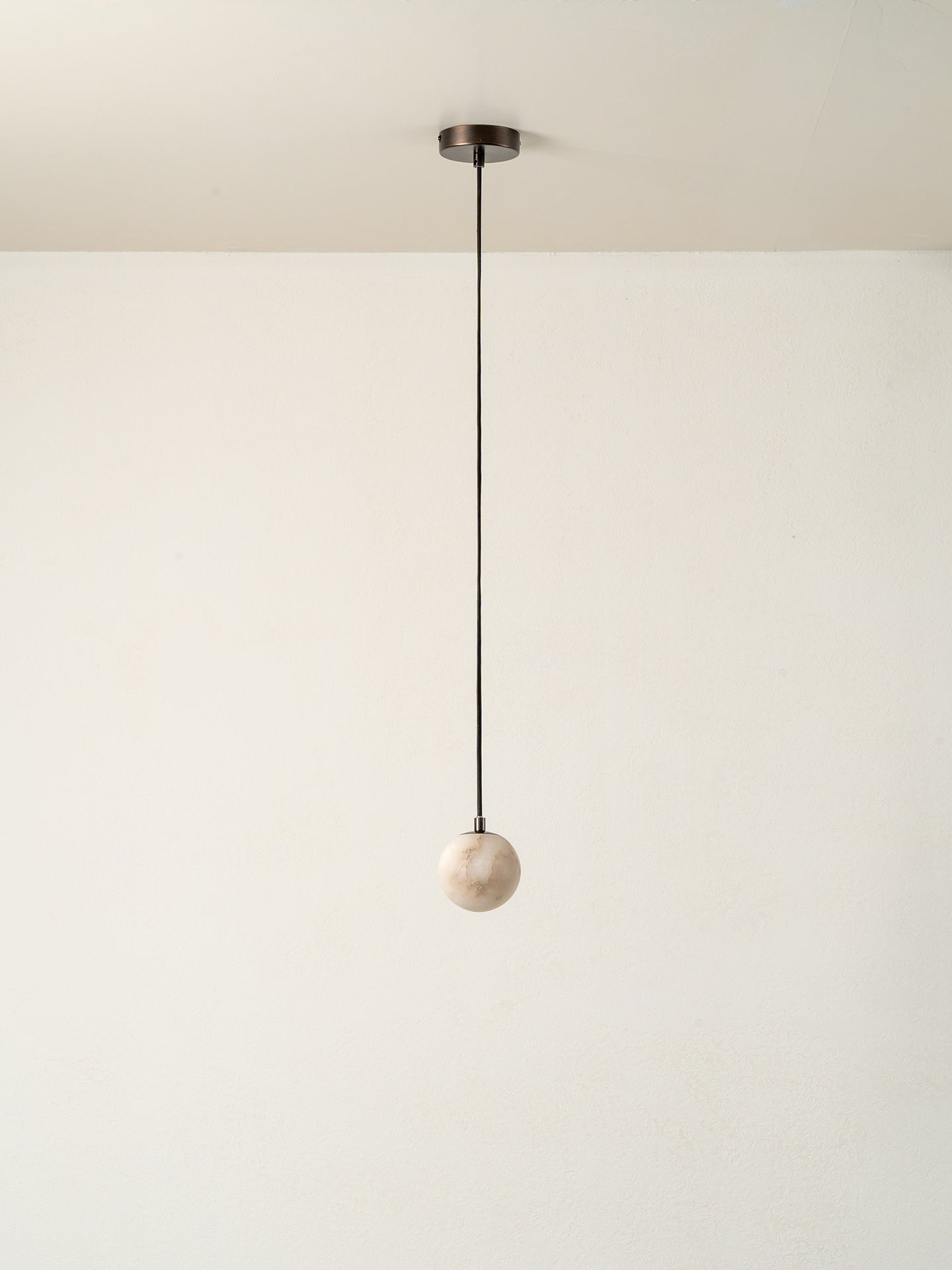 A minimalist pendant light with a matte black cord and ceiling canopy, suspending a small, round, textured off-white marble or stone globe shade against a plain off-white wall.