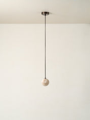 A minimalist pendant light with a matte black cord and ceiling canopy, suspending a small, round, textured off-white marble or stone globe shade against a plain off-white wall.