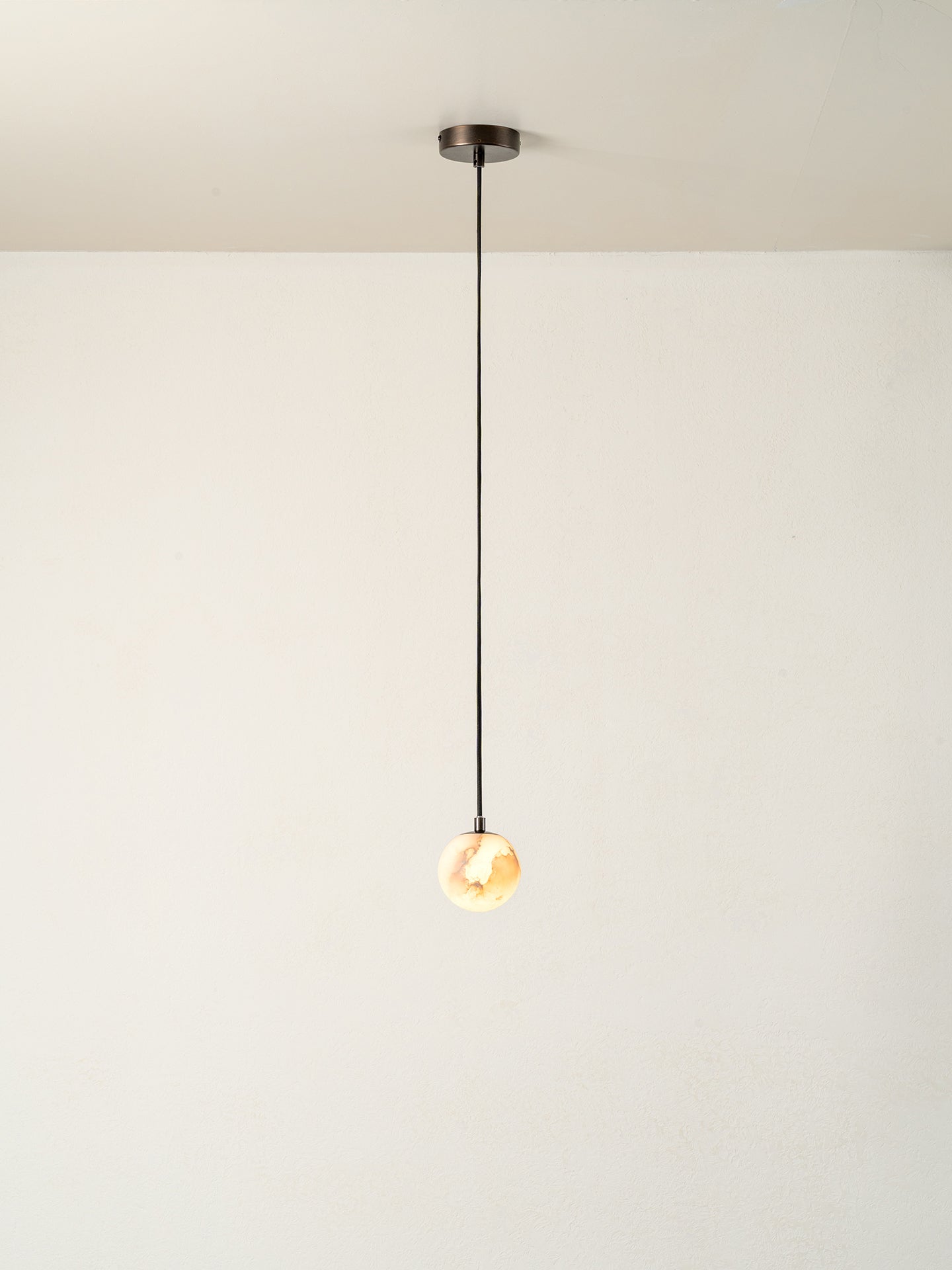 A minimalist single-light pendant ceiling fixture with a dark metal canopy, a thin black cord, and a glowing warm glass globe bulb, shown against a plain off-white wall and ceiling.
