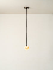 A minimalist single-light pendant ceiling fixture with a dark metal canopy, a thin black cord, and a glowing warm glass globe bulb, shown against a plain off-white wall and ceiling.
