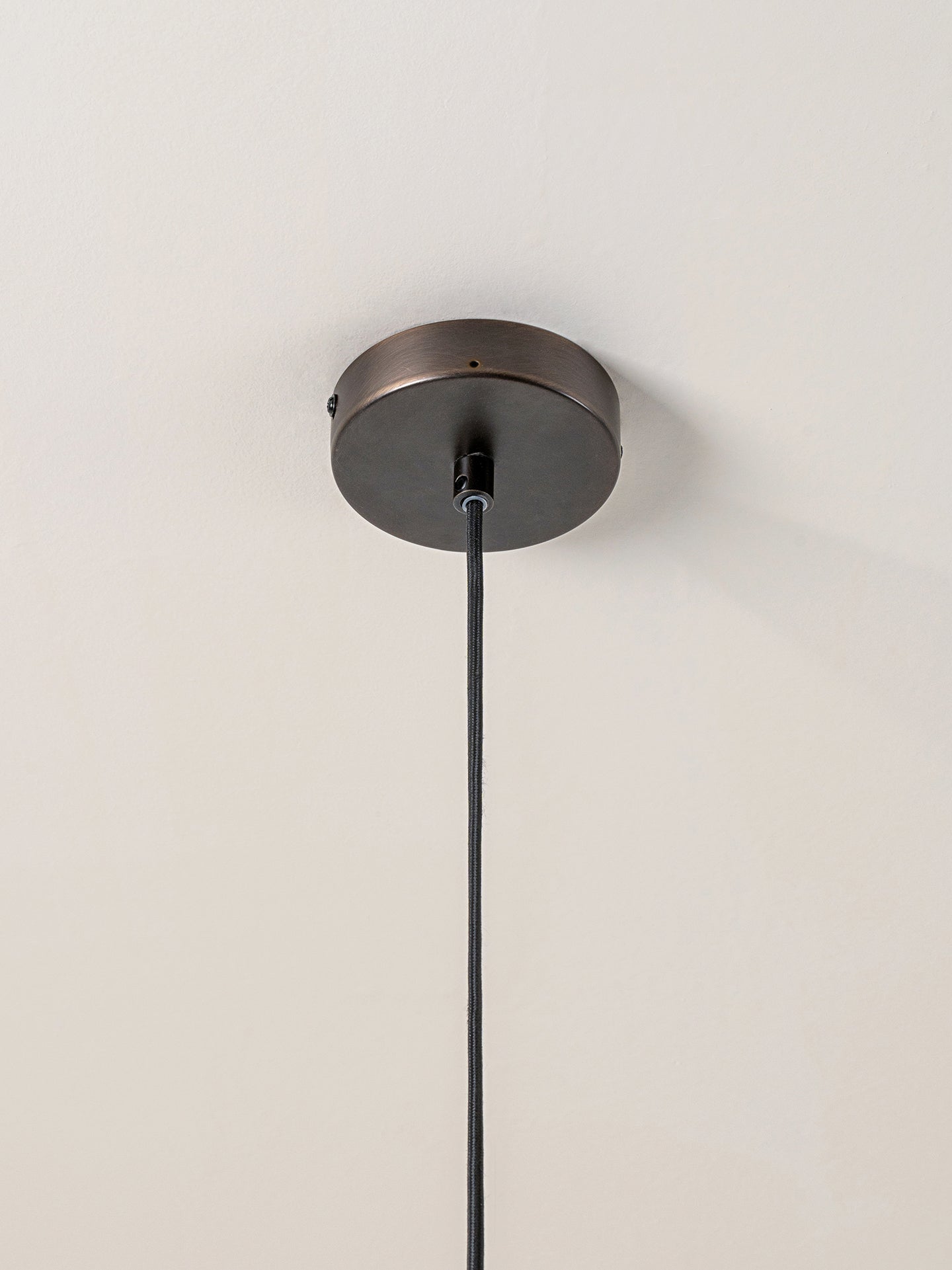 A close-up of a ceiling-mounted light fixture showing a round dark bronze ceiling canopy with a black hanging cord against a plain off-white wall.