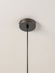 A close-up of a ceiling-mounted light fixture showing a round dark bronze ceiling canopy with a black hanging cord against a plain off-white wall.
