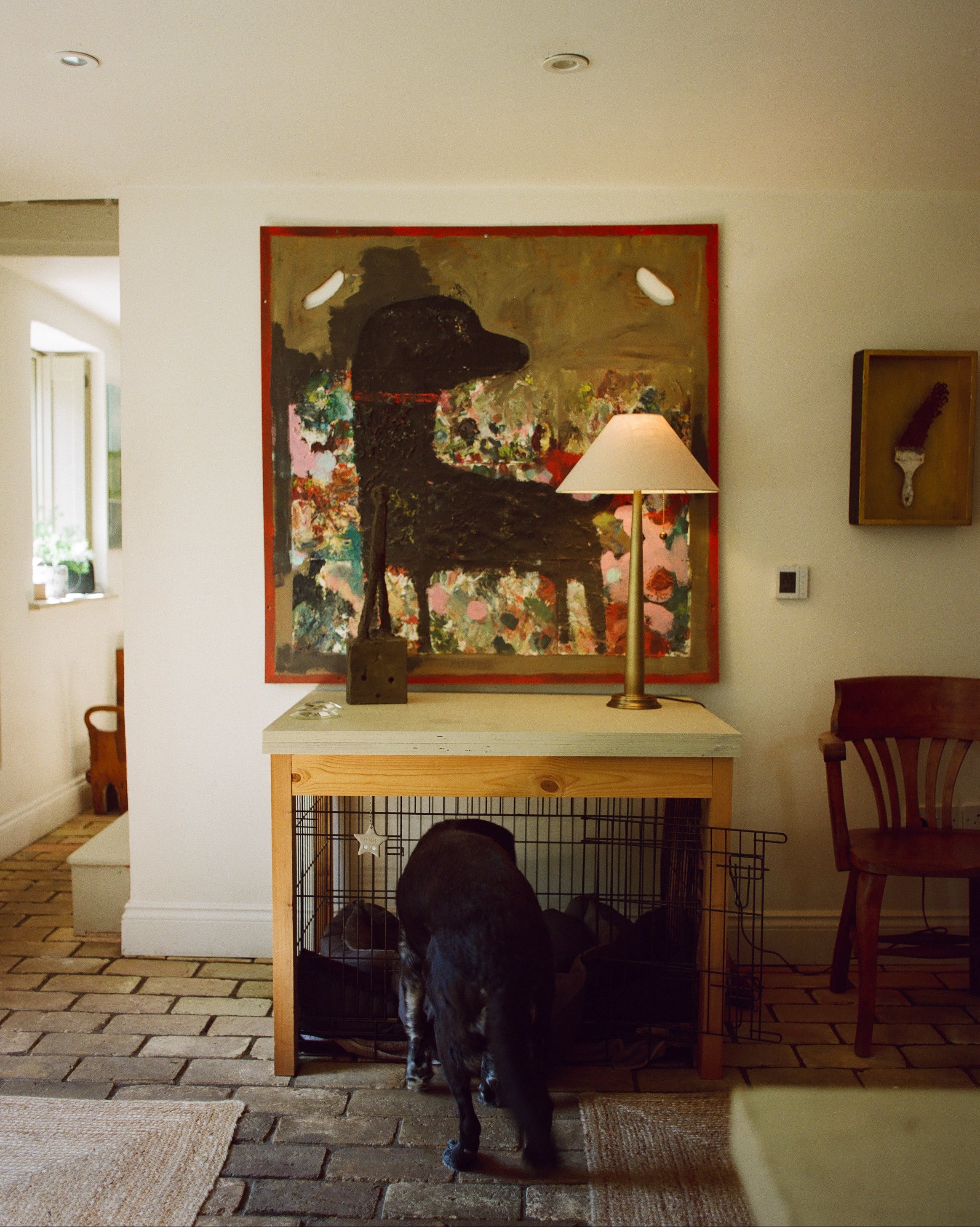 A large framed painting of a black dog hangs on a cream wall above a wooden console table in a home interior. A black dog stands in front of the table, and a lamp sits on the tabletop.