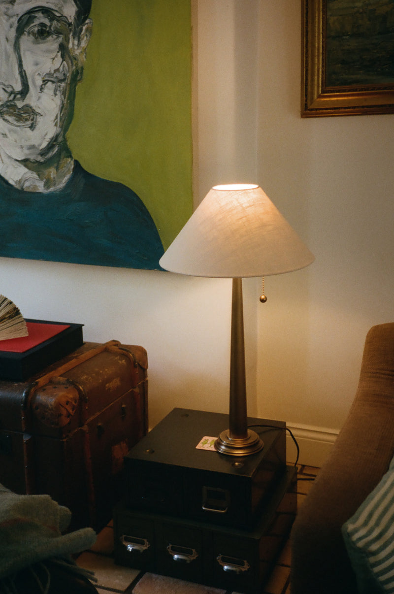 A table lamp with a warm white fabric shade and a dark metallic base sits on a black equipment case in a cozy room corner, next to a brown leather chair and a vintage suitcase under a large colorful portrait painting.