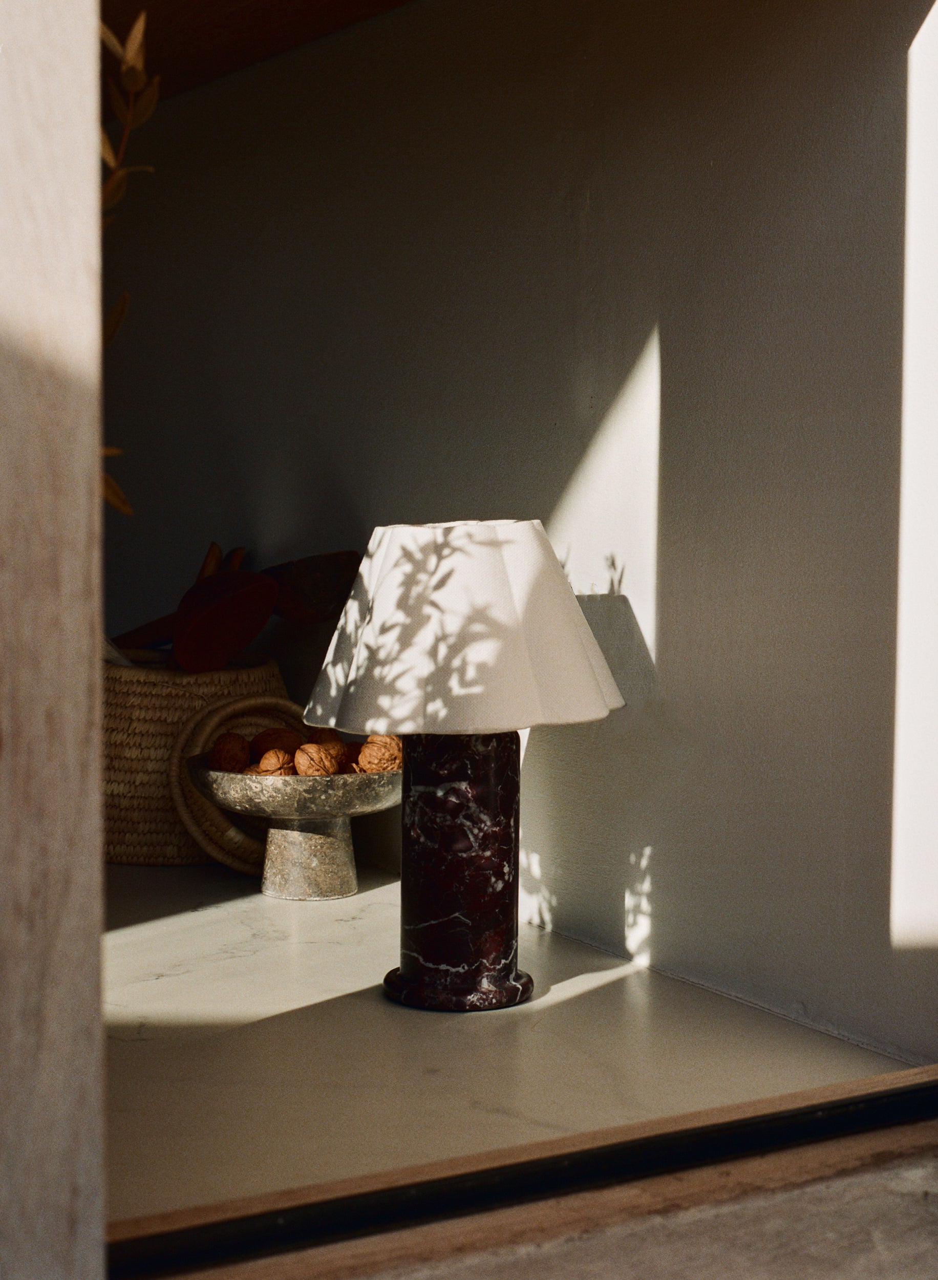 A table lamp with a white tapered shade and a dark polished marble base sits on a shelf in a sunlit interior, with dappled shadows cast across the shade and wall.