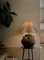 A rustic ceramic table lamp with a dark brown clay body and a pleated beige fabric shade, illuminated and displayed on a wooden stool in a warm, modern interior setting.