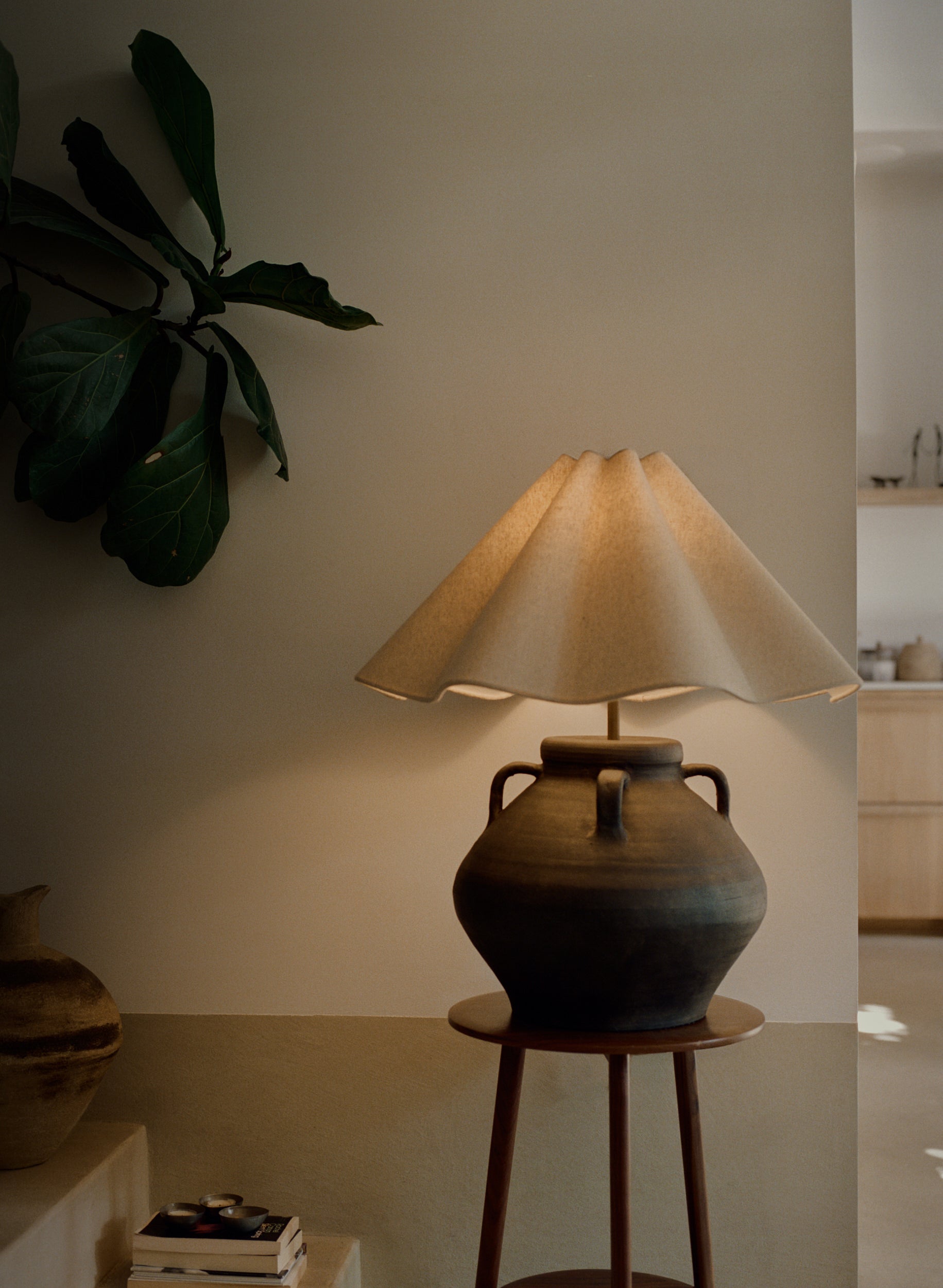 A rustic ceramic table lamp with a dark brown clay body and a pleated beige fabric shade, illuminated and displayed on a wooden stool in a warm, modern interior setting.