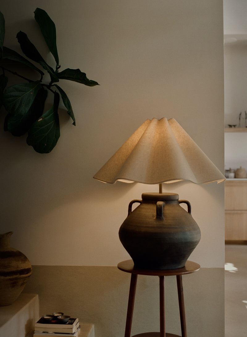 A rustic ceramic table lamp with a dark brown clay body and a pleated beige fabric shade, illuminated and displayed on a wooden stool in a warm, modern interior setting.