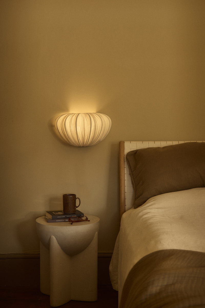 A modern wall-mounted ribbed pendant light glowing warmly on a beige wall beside a bed with cream bedding and a small sculptural side table.