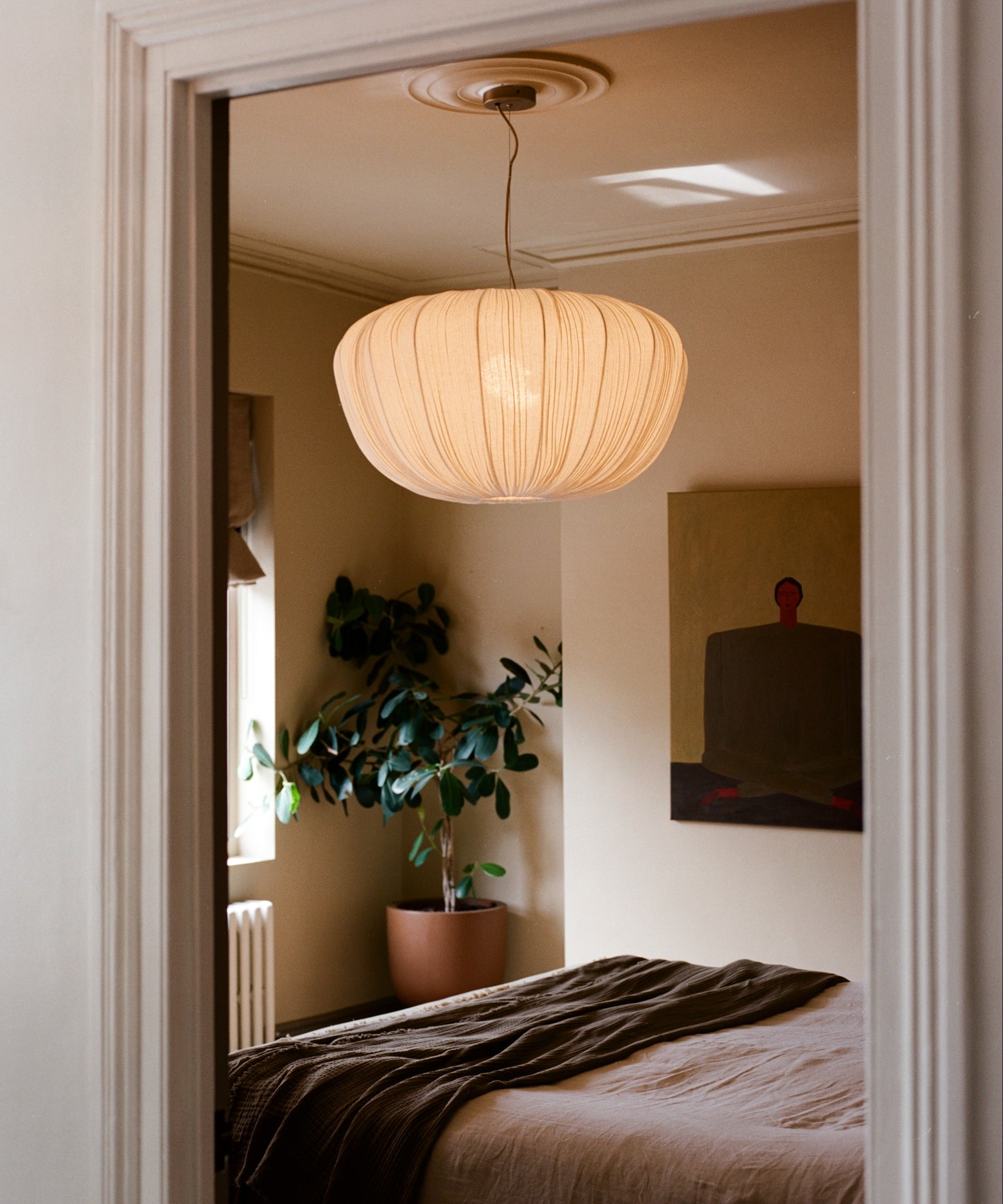 A modern pleated pendant light in a warm cream color hangs in a softly lit bedroom, viewed through an open doorway. The room features neutral beige walls, a bed with brown bedding, and a potted green plant in the corner.