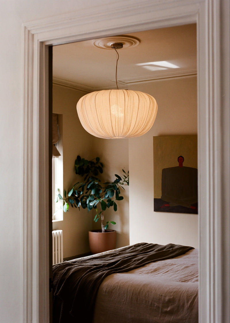 A modern pleated pendant light in a warm cream color hangs in a softly lit bedroom, viewed through an open doorway. The room features neutral beige walls, a bed with brown bedding, and a potted green plant in the corner.