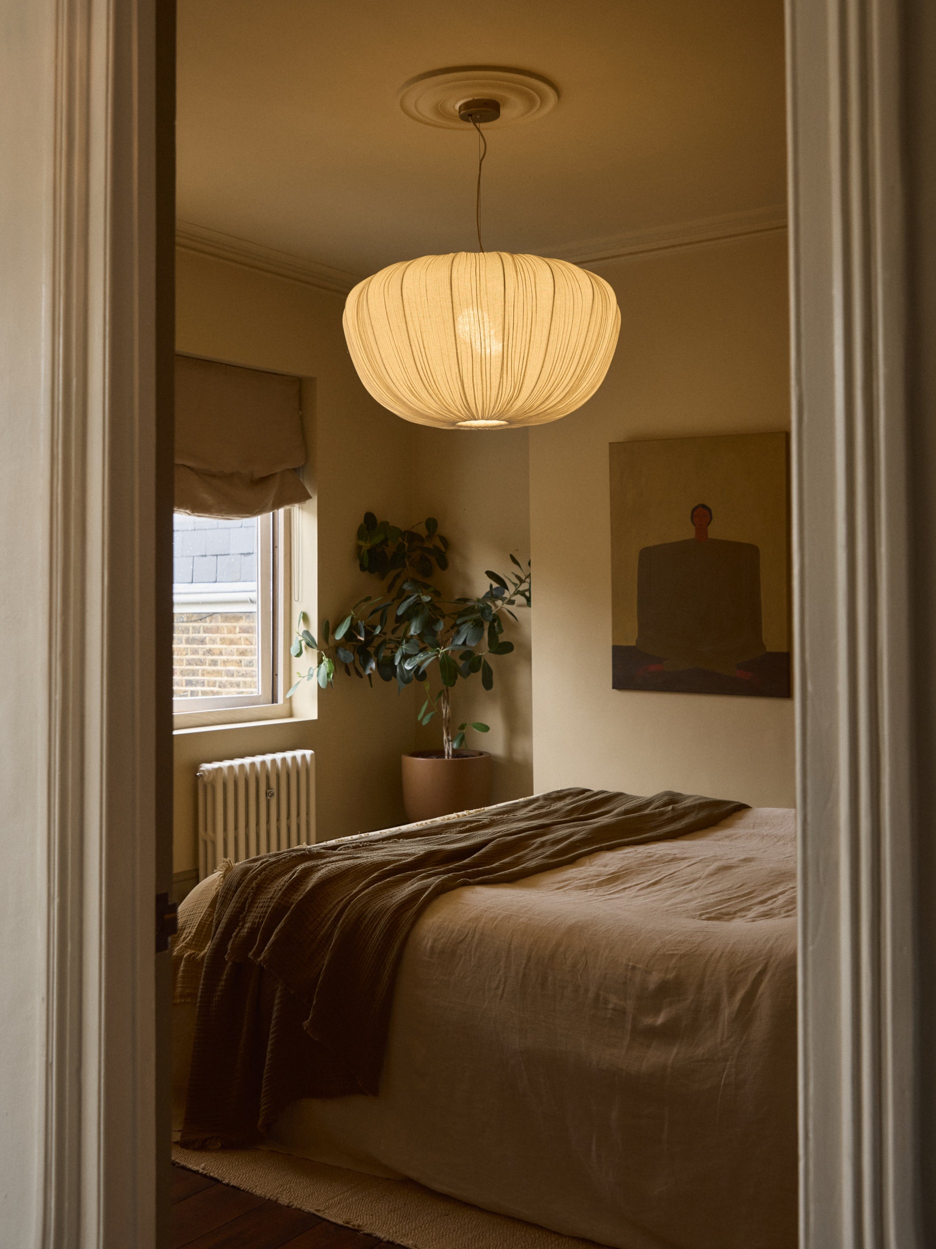 A warm, modern bedroom scene viewed through an open doorway, featuring a large pleated pendant light hanging above a bed with beige linens and a brown throw blanket.