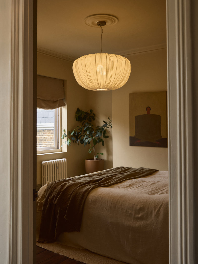 A warm, modern bedroom scene viewed through an open doorway, featuring a large pleated pendant light hanging above a bed with beige linens and a brown throw blanket.