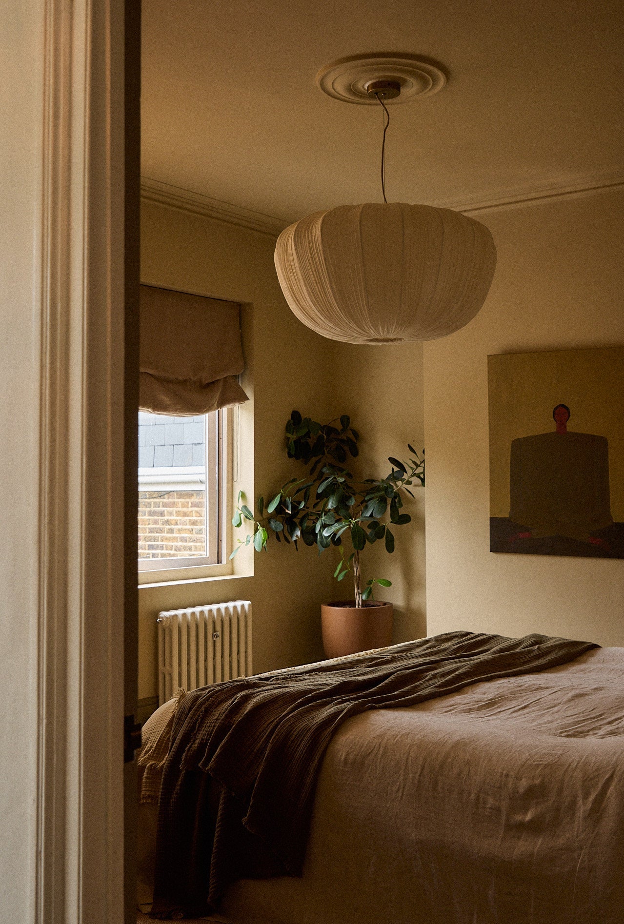 A warm, minimalist bedroom scene viewed through an open doorway, featuring a bed with beige linens and a dark brown throw blanket, a large white pleated pendant light, a potted green plant, and a painting on the wall.