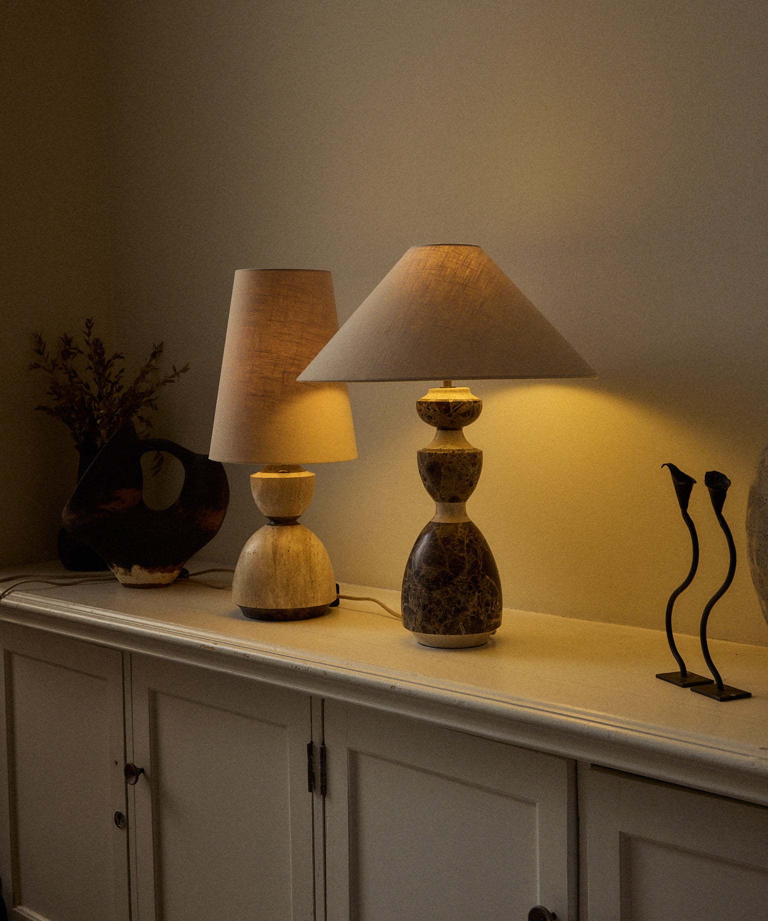 Two table lamps displayed on a white sideboard in a warmly lit room. The lamp in the foreground has a tiered stone base in earthy brown and beige tones with a beige conical shade.