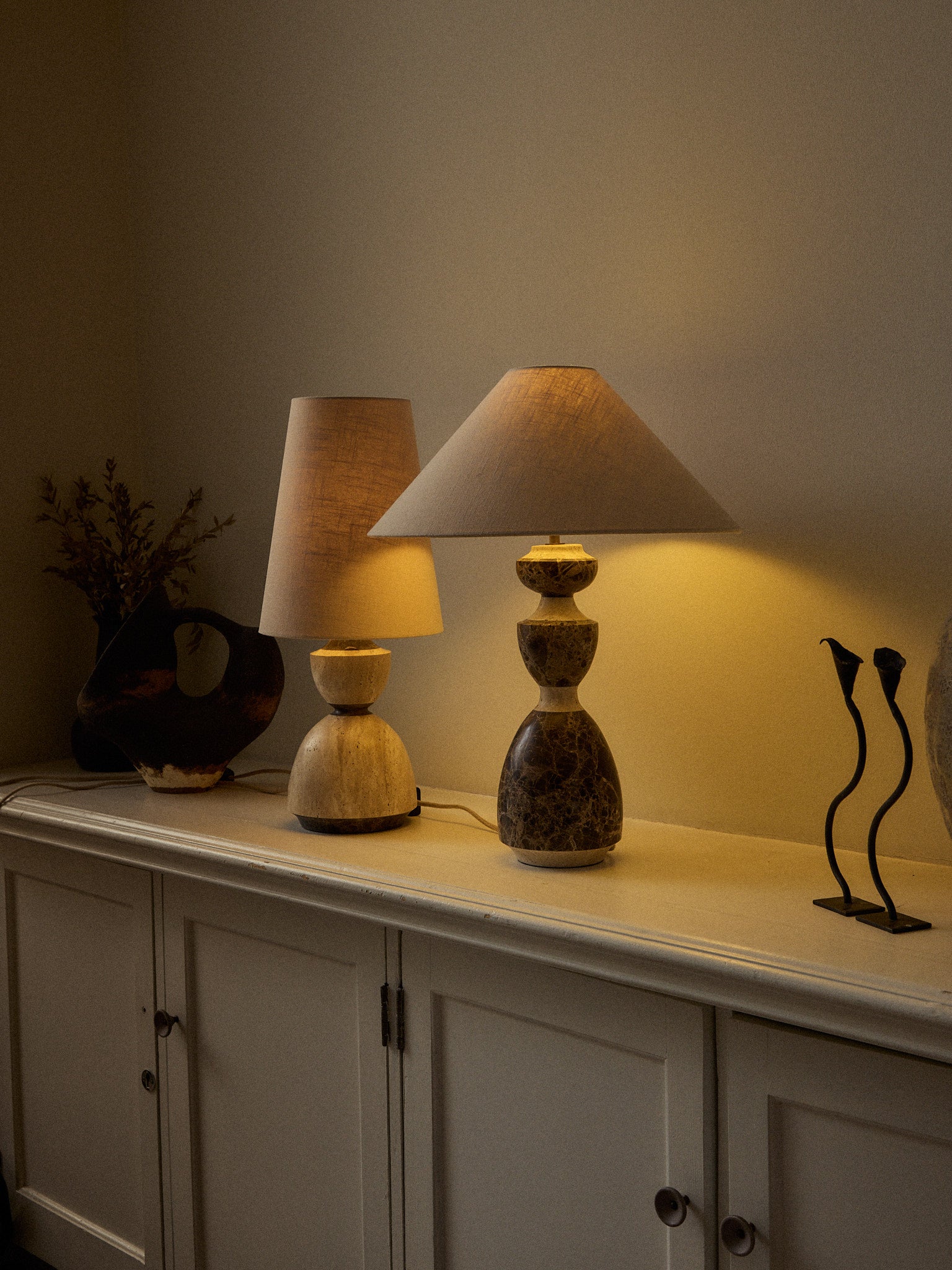 Two table lamps displayed on a white sideboard in a warmly lit room. The lamp in the foreground has a tiered stone base in earthy brown and beige tones with a beige conical shade.