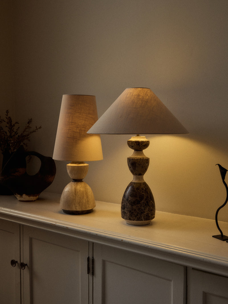 Two table lamps displayed on a cream-colored cabinet in a warm, dimly lit room. The lamp on the right features a dark brown and beige marbled base with a beige fabric drum shade, while the lamp on the left has a lighter beige stone base and matching shade.