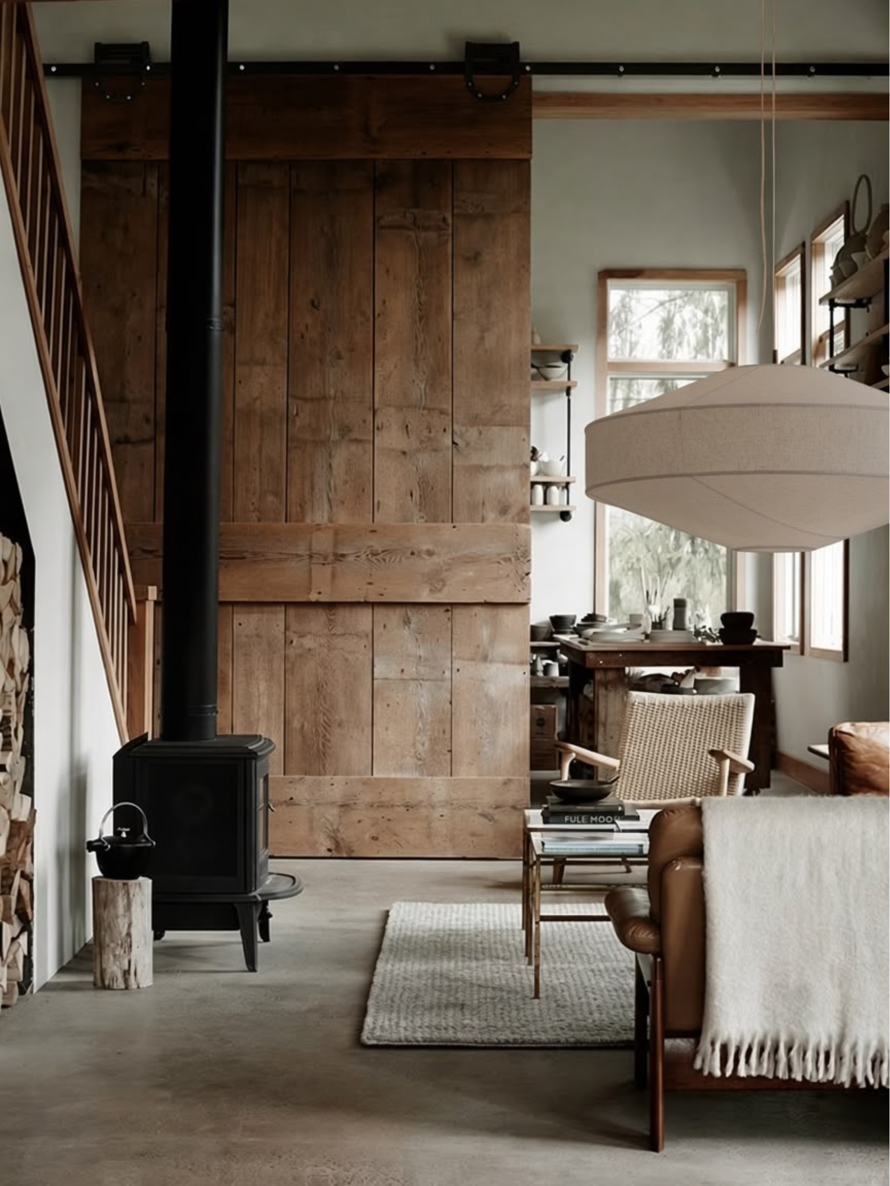 A rustic interior scene with a large wooden barn-style door, black wood-burning stove, staircase, dining table, pendant light, and seating area with a white throw blanket.