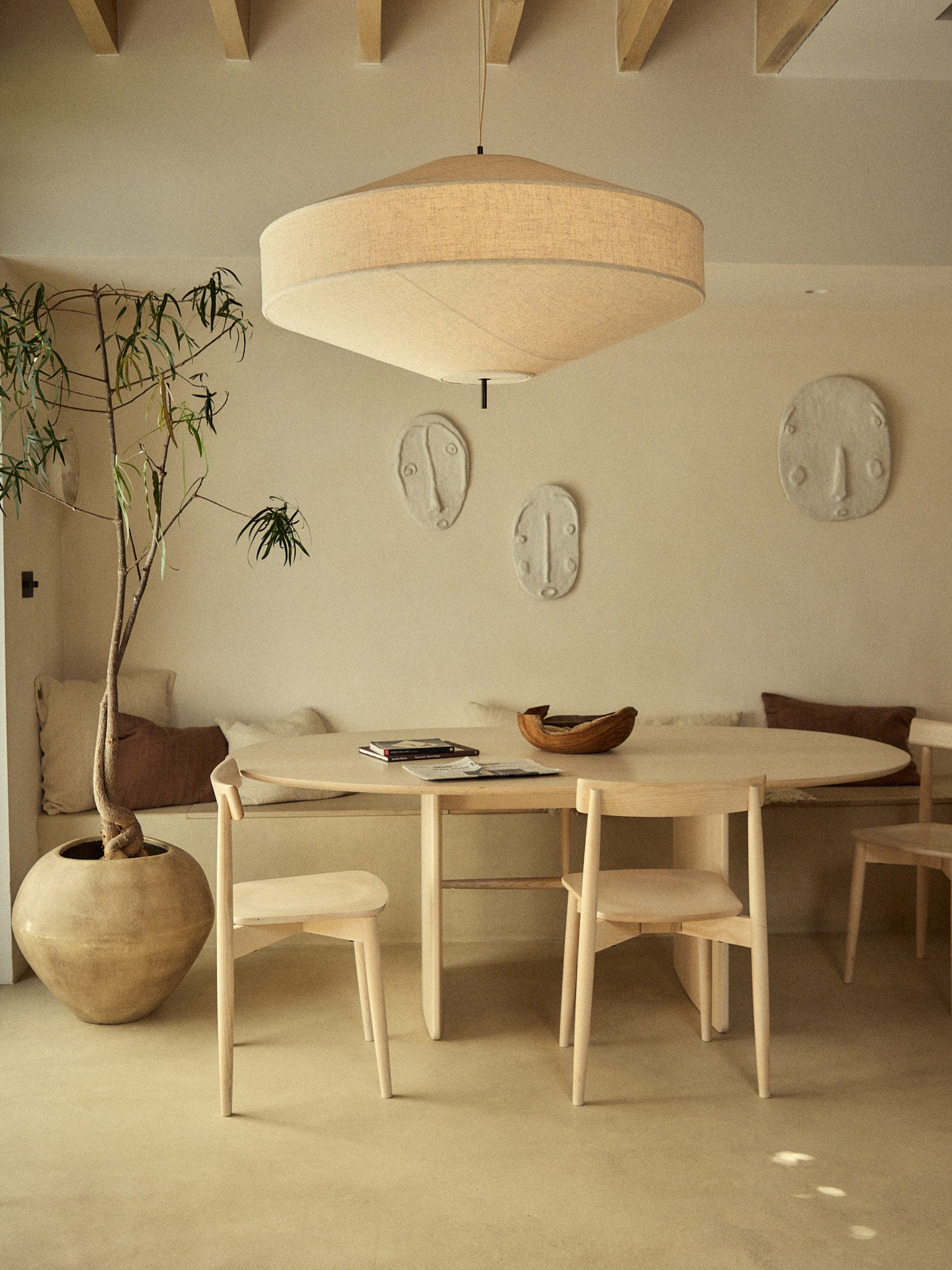 A minimalist dining area featuring a light wood oval table, four matching wooden chairs, a large cylindrical beige pendant light, and a tall potted plant in a textured ceramic pot against a warm beige plaster wall.