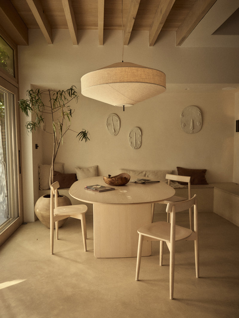 A minimalist dining area featuring a round light wood table, three matching light wood chairs, a large drum-shaped pendant lamp, and a built-in bench with cushions against a warm beige wall.