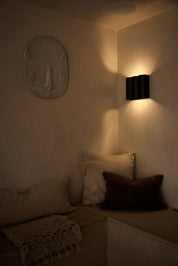 A modern black wall sconce with a vertical pleated design, mounted on a beige wall in a dimly lit room corner above a low bench with neutral-toned cushions.