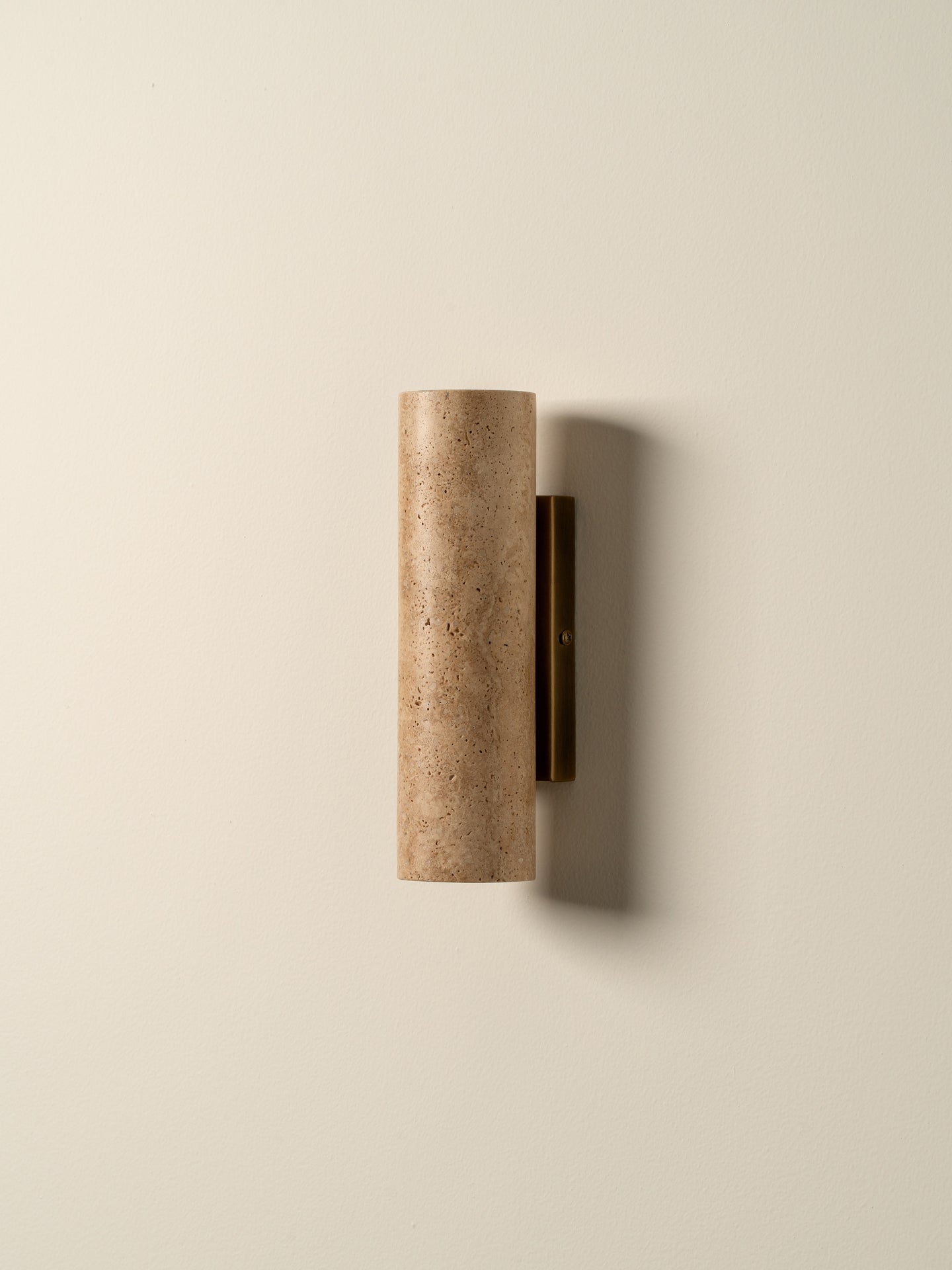 A modern cylindrical wall sconce with a textured beige travertine stone shade and a dark bronze vertical backplate, mounted on a warm off-white wall.