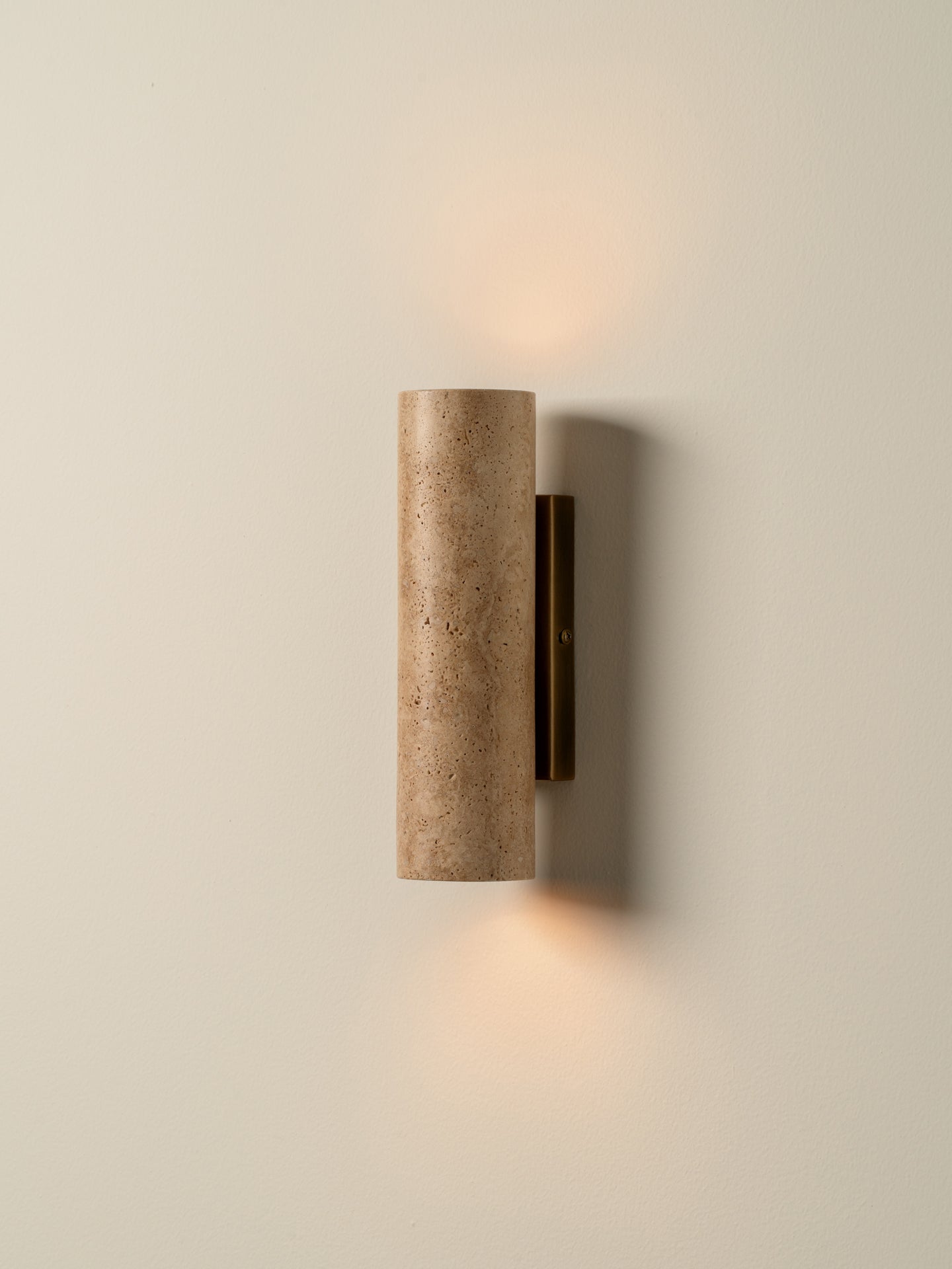 A modern cylindrical wall sconce with a textured beige travertine stone shade and a dark bronze metal mounting bracket, shown illuminated against a warm off-white wall.